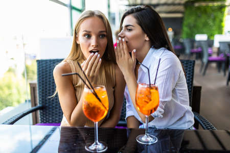 Two women sharing secrets and covering faces while drink cocktails in cafe.の写真素材
