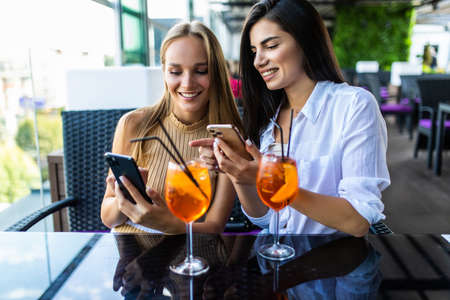 Two women smiling and using cellphones while drinking cocktails at table in cafe outdoorsの写真素材