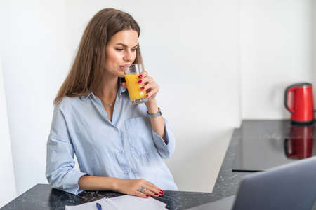 Woman using a laptop while drinking juice in her kitchen working from home in quarantine lockdown.