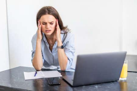 Young tired woman in casualwear sitting in front of laptop while touching her forehead during work...
