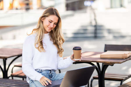 selective focus of beautiful girl in casual clothes holding paper coffee cup while sitting on wooden bench in park and using laptopの写真素材