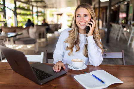 Portrait of young business woman with glasses sitting in cafe in front of her laptop and talking on mobile phoneの写真素材