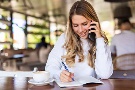 Cheerful young woman laughing during phone conversation on smartphone doing order and making notes...