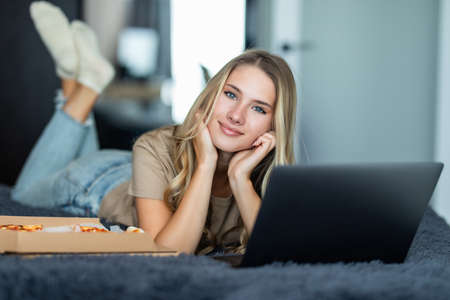 Young woman lying on a bed in bed at home while eating pizza and using laptopの写真素材