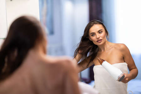 beautiful young woman holding hair brush and drying hair at mirror in bathroom