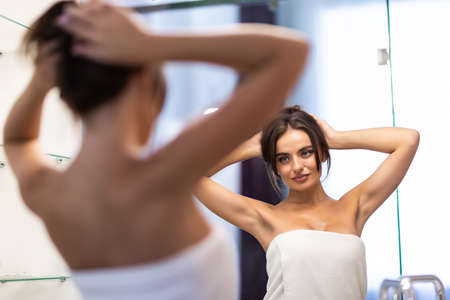 beautiful smiling young woman in bathrobe and towel on head looking at mirror in bathroomの写真素材