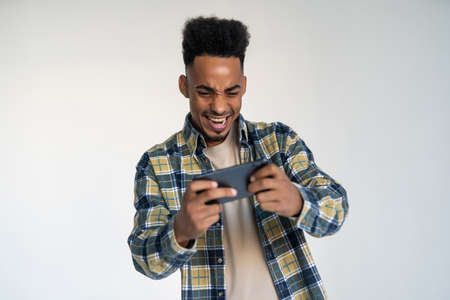 Portrait of a young african man dressed in plaid shirt playing games on mobile phone isolated over...