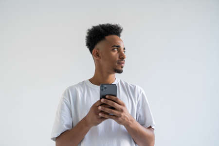 Daylight portrait of young african american man isolated on gray background wearing white t-shirt standing in front of camera, looking attentively with smile at screen of smartphone he is holdingの写真素材