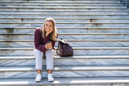 Portrait of an attractive happy woman holding coffee cup while sitting on stairs at the city streetの写真素材