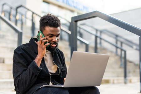 Handsome African-American man in casual clothes working with a laptop and talking on smart phone while sitting on stairsの写真素材