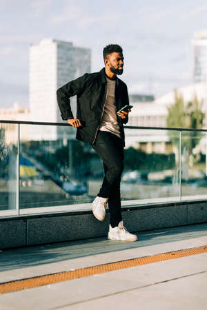 Young Afro American man with coffee cup standing smiling and use on cell phone in the streetの写真素材