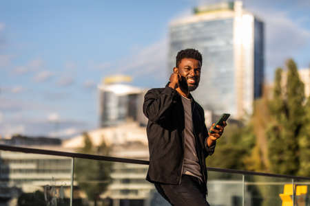 Happy young African American man in headphone walking at sunny city and making video conversation...