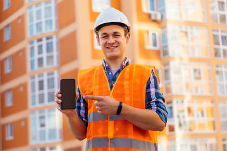 Architecture, building business and people concept. Smiling male architect in helmet showing phone over construction site backgroundの写真素材