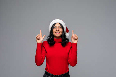 Pretty brunette girl in a red sweater and Santa hat pointing at the copy space up isolated over gray...