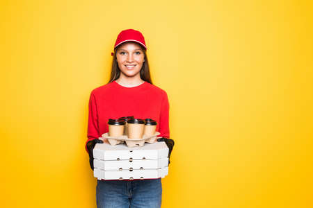 Delivery employee woman in red cap blank t-shirt uniform work courier service hold food order pizza in cardboard flatbox, paper cups of coffee tea on yellow backgroundの写真素材