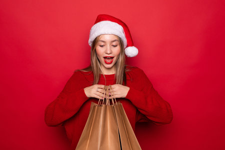 Surprised young Santa woman wearing Christmas hat hold package bags with purchases after shopping isolated on red background studio portraitの写真素材