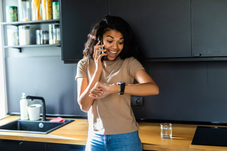 Young pretty woman tooking at her wrist watch while sitting on a table in a kitchenの写真素材