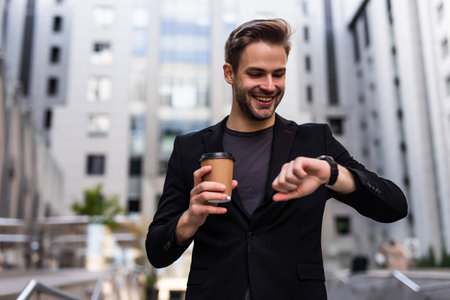 Confident male entrepreneur checking time on watch during break while hurrying for meeting, serious businessman in formal suit standing on urban settings with coffee takeaway looking at wristwatchの写真素材