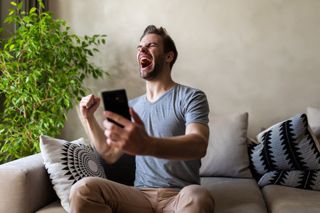 Smiling young man holding mobile phone while sitting on a sofa at home with laptop computer and celebratingの写真素材