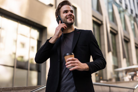Attractive confident young businessman wearing a suit sitting on a bench outdoors at the city street, talking on mobile phone while drinking takeaway coffeeの写真素材