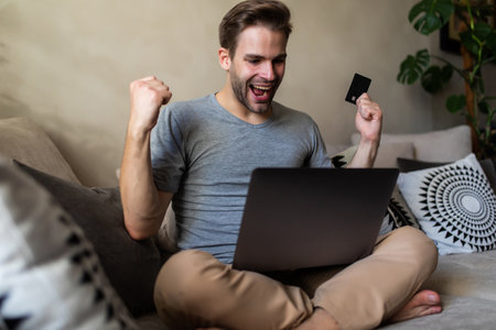 happy excited young man with laptop at home sitting on a couchの写真素材