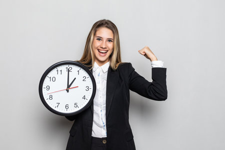 Portrait of beautiful young business woman holding in hands clock with win gesture, isolated on...