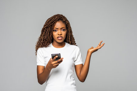 Portrait of confused puzzled woman with afro hairstyle wearing t-shirt holding cell phone and...