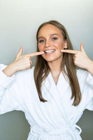 Beautiful girl in bathrobe and with towel on her head is looking at camera, pointing and smiling, standing in bathroomの写真素材