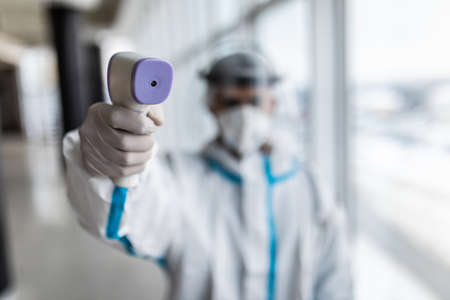 Medical worker scanning with infrared thermometer, IR Thermoscan. Confident Asian doctor in protective PPE suit wearing face mask and eyeglasses in hospitalの写真素材