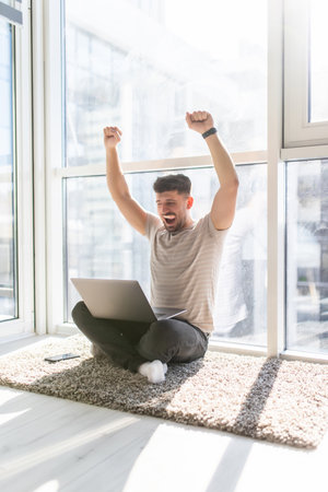 Excited young man working gesturing and looking at laptop while sitting on the floor at his apartmentの写真素材
