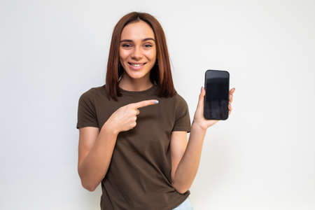 Cheerful cute woman pointing finger on smartphone screen isolated on a white background. Looking at...