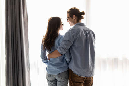Back view of beautiful young couple looking out the window and hugging while having a breakfast at homeの写真素材