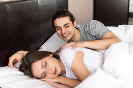 Happy handsome young man lying and waking his woman in bed