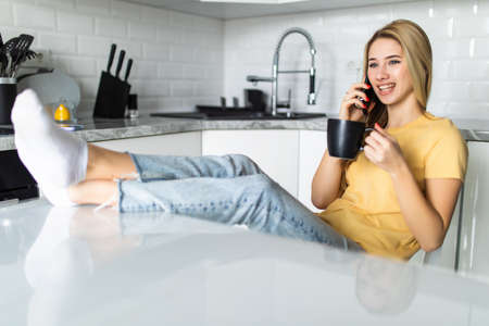 portrait of young beautiful woman talking cell phone, drink coffee or tea in the kitchenの写真素材