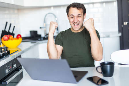Excited man sit at table in kitchen feel euphoric win online lottery on laptop.の写真素材