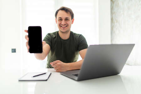 Happy handsome man freelancer sitting at the kitchen table, working on laptop computer, showing blank screen mobile phoneの写真素材