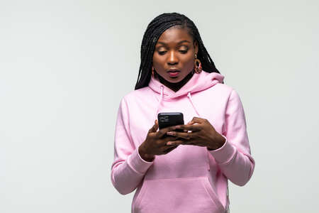 Shocked young african american woman using mobile cell phone typing sms message on white background. People lifestyle concept.の写真素材