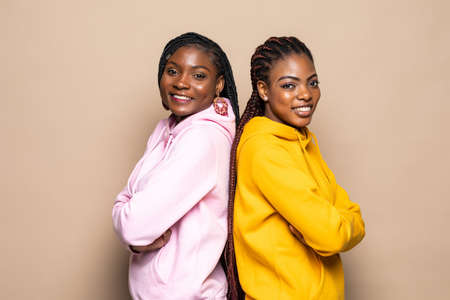 Portrait of two cheerful young women standing together and looking at camera isolated over beige backgroundの写真素材