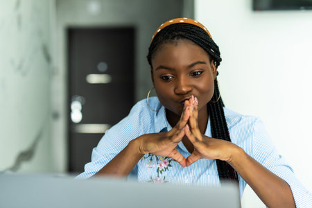 Attractive young african woman on a video call with colleagues, sitting on a kitchen with laptop.