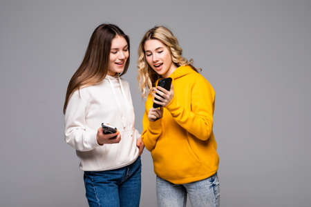 Two cheerful excited young women use smartphones over gray background