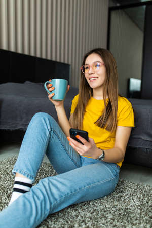 young woman use the smartphone and drink coffee, sitting on the floor in living roomの写真素材