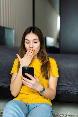 Surprised young woman using smartphone while sitting on floor at home