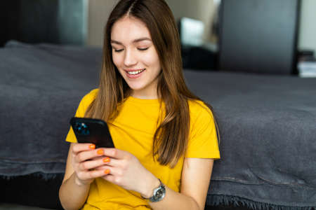 White young woman smiling and using mobile phone while sitting on couch at homeの写真素材