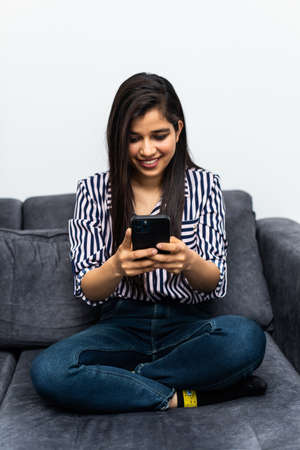 Happy indian woman relaxing on comfortable couch, holding smartphone in hands. Smiling young lady chatting in social networks, watching funny videos, using mobile applications at home.の写真素材