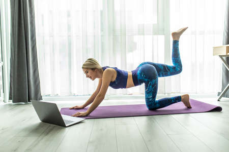 Gorgeous young woman sitting on exercise mat at home and watching workout tutorial online on laptop....