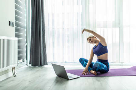 Hatha yoga concept. Concentrated woman stretching hands on yoga mat, sitting near laptop computer,...