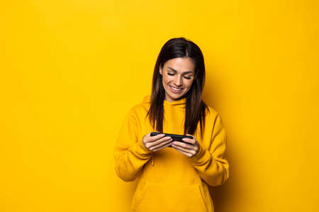 Happy beautiful young woman play games by mobile phone isolated over yellow wall background.
