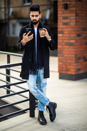 Young indian man use phone holding coffee to go while walking on the street