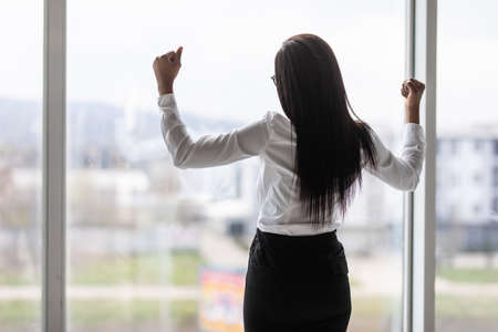 African businesswoman spreading hands standing at office window, enjoying big city, successful entrepreneur celebrating business success with arms open wide, feeling powerful inspired, rear viewの写真素材