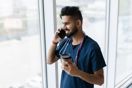 Young Indian doctor talk phone in hospital. Consulting doctor. Medical staff in clinic. Bearded man with smartphone. Healthcare Professional Concept.の写真素材